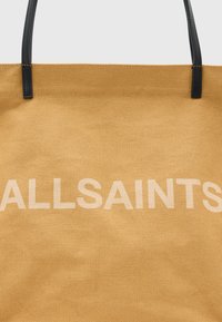 Tote bag in mosterdgeel, gemaakt van textuurstof, met zwarte leren handvatten en de merknaam "ALLSAINTS" in het wit prominent weergegeven.