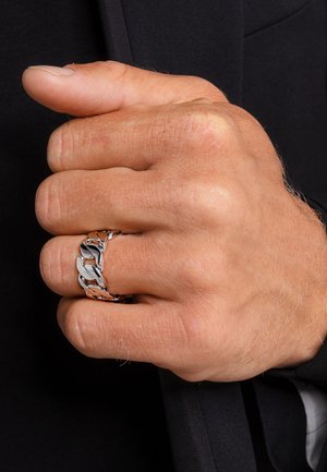 Mano maschile chiusa a pugno con un grosso anello a maglia di catena in argento al dito medio su uno sfondo di tessuto nero.