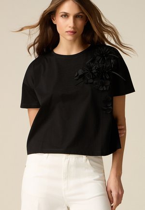 Donna con capelli lunghi che indossa una maglietta nera a maniche corte con decorazioni floreali 3D sulla spalla, abbinata a pantaloni bianchi.