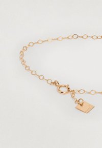 Arany színű karkötő, gyémánt alakú láncszemekkel, homárkapoccsal és egy kis taggel, amelyen gravírozott szöveg található. Simának tűnő felület, finom dizájn.