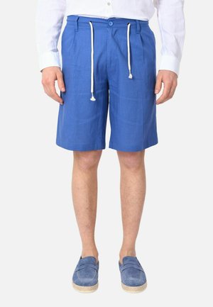 Uomo che indossa pantaloncini blu con coulisse, camicia bianca con bottoni e scarpe blu slip-on, in piedi davanti a uno sfondo bianco.
