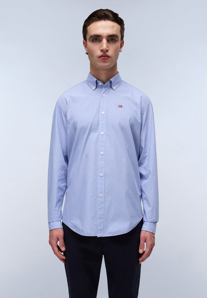 Light blue, long-sleeve shirt with white vertical stripes, buttoned collar, and small branded patch on the chest. Flat buttons and curved hem.
