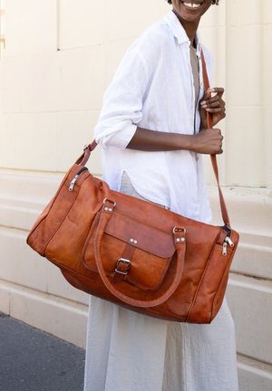 Personne en chemise blanche et pantalon portant un grand sac de voyage en cuir marron avec plusieurs poches et une bandoulière.