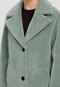 Veste en polaire verte avec un col cranté, des boutons noirs, et une surface texturée. Portée sur une chemise noire.