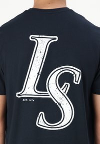 Tamsiai mėlynas medvilnės marškinėlis su dideliu baltu nusidėvėjusiu „LS“ grafiniu ženklu ir „EST. 1874“ tekstu nugarėlėje. Trumpomis rankovėmis ir apvaliu kaklu.