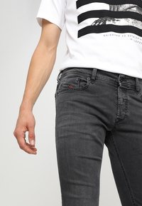 Jeans de mezclilla negra de corte ajustado, diseño de cinco bolsillos y sutil detalle del logo en rojo. Combinados con una camiseta blanca con un estampado gráfico.