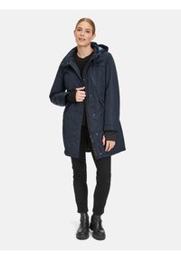 Marineblaue wasserdichte Parka mit Kapuze, elastischem Stoff, Reißverschluss und Druckknöpfen sowie seitlichen Taschen; getragen über schwarzen Hosen und Stiefeln.
