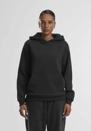 Frau steht aufrecht, trägt einen schlichten schwarzen Hoodie mit Kängurutasche und dunkle Hose vor einem schlichten grauen Hintergrund.