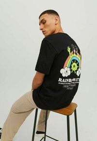 Must musta T-särk, mille seljal on värviline vikerkaare graafika, kus on naeratav päike ja pilved. Kandmine beežide pükste ja mustade kingadega.