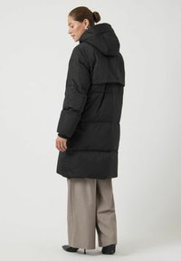 Giacca a piumino nera con cappuccio, caratterizzata da un taglio squadrato, motivo trapuntato e maniche lunghe. Indossata sopra pantaloni beige a gamba larga.