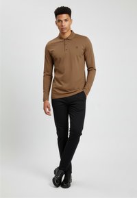 Junger Mann steht mit den Händen in den Taschen, trägt ein braunes langärmeliges Poloshirt, schwarze Hose und schwarze Lederschuhe vor einem weißen Hintergrund.