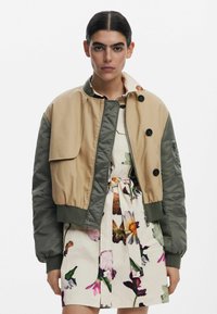 Khaki en olijfgroene bomberjack met ritssluiting aan de voorkant, geribde manchetten en een oversized voorzak. Draagt boven een bloemenjurk.