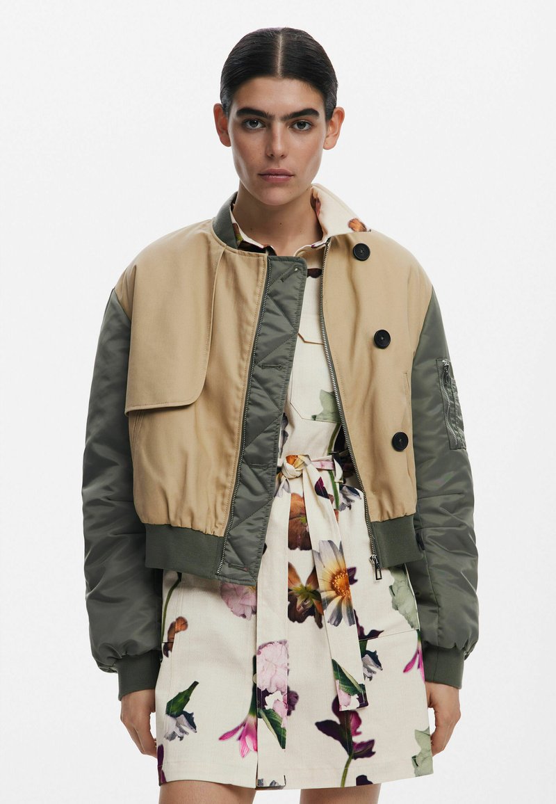 Khaki en olijfgroene bomberjack met ritssluiting aan de voorkant, geribde manchetten en een oversized voorzak. Draagt boven een bloemenjurk.