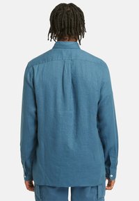 Camisa de linho azul de mangas longas com colarinho abotoado, detalhe de pregas nas costas e punhos com um botão. Textura suave e ajuste descontraído.