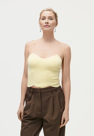 Femme portant un haut léger jaune pâle à fines bretelles avec des détails noués et un pantalon marron plissé taille haute, debout devant un fond uni.