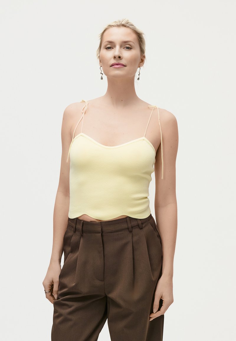 Γυναίκα που φοράει ανοιχτό κίτρινο τοπ με λεπτές τιράντες και ζώνη με κόμπο, συνοδευόμενο από καφέ ψηλόμεσο πλισέ παντελόνι, στέκεται μπροστά σε απλό φόντο.