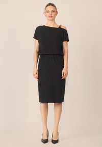 Schwarzes Kleid mit kurzen Ärmeln und asymmetrischem Ausschnitt. Verfügt über eine taillierte Taille und einen knielangen Rock. Enthält ein dekoratives Schulterdetail.