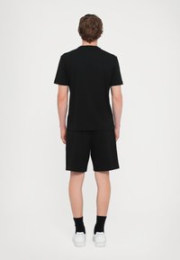 Black cotton T-shirt with short sleeves, paired with black shorts, viewed from the back. White sneakers and black socks complete the look.