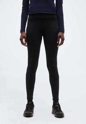 Osoba nosiaca čierne priliehavé nohavice, čierne športové topánky a tmavomodrú košeľu s dlhým rukávom stojaca na bielom pozadí.