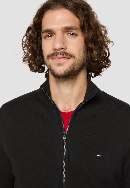 Schwarze Fleecejacke mit Reißverschluss und hohem Kragen, aus strukturiertem Material, mit einem kleinen rot-weißen-blauen Logo auf der Brust.