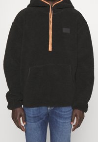 Pullover in pile nero con zip arancione, grande tasca frontale e tessuto testurizzato. Presenta un cappuccio e una piccola etichetta con logo nero.