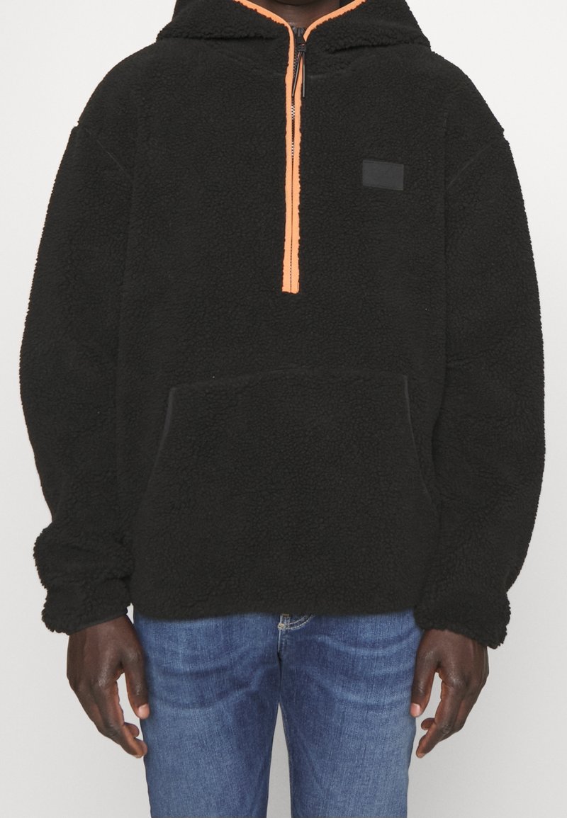 Pullover in pile nero con zip arancione, grande tasca frontale e tessuto testurizzato. Presenta un cappuccio e una piccola etichetta con logo nero.