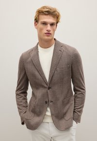 Brauner kariierter Blazer mit zwei Knöpfen, Reverskragen und aufgesetzten Taschen, getragen über einem hellen Pullover und kombiniert mit weißen Hosen.