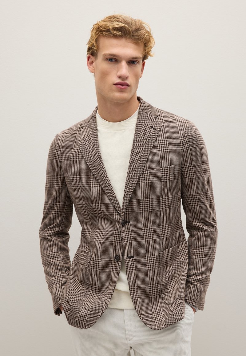 Brauner kariierter Blazer mit zwei Knöpfen, Reverskragen und aufgesetzten Taschen, getragen über einem hellen Pullover und kombiniert mit weißen Hosen.