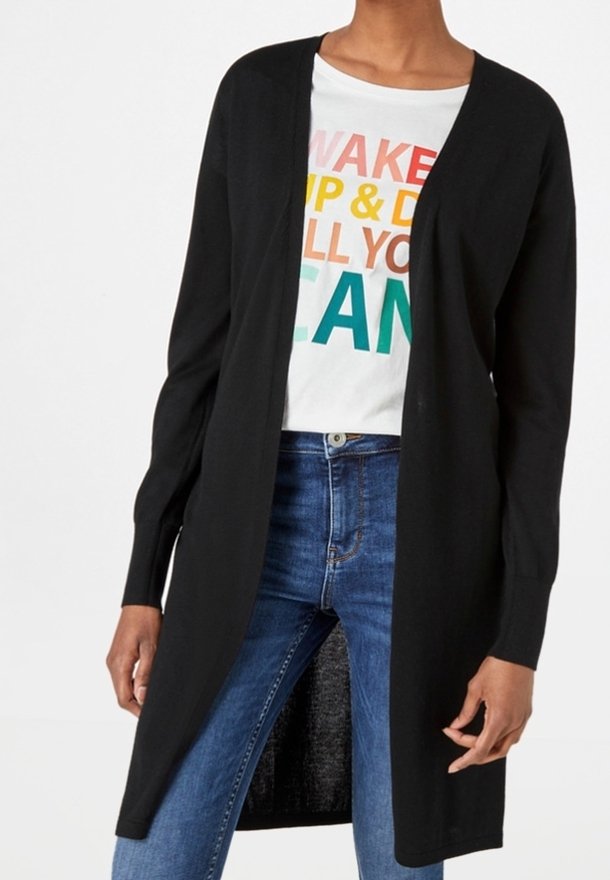 Henkilö, joka käyttää pitkää mustaa avointa kardigania valkoisen paidan, jossa on värikkäitä tekstejä, ja sinisiä farkkuja, seisoo rentoina käsivarret vartalon sivuilla.