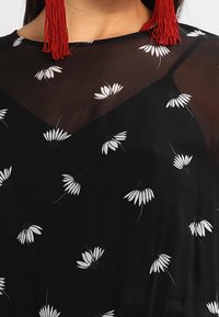 Top negro con una capa de tela transparente con un patrón floral en blanco. Pendientes de borlas rojas añaden contraste y detalle.
