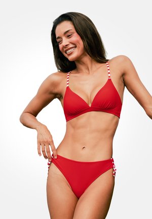 Mujer sonriendo y posando con un bikini rojo con tirantes a rayas y lazos laterales sobre un fondo blanco liso.