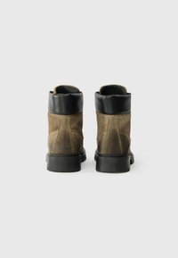 Botas de tornozelo em camurça castanha com um colarinho de couro preto, bico redondo e sola exterior de borracha preta sólida, apresentando detalhes de costura minimalistas.