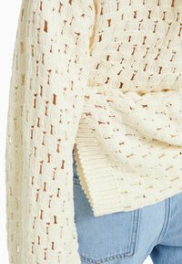 Krämfärgad stickad tröja med en strukturerad öppen väv och ribbade detaljer på ärmsluten och nederkanten. Parat med bl åjeans.