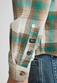 Geruit flanellen overhemd met bruin, petrol en wit; zachte textuur, knoopmanchetten en een geweven label met het "Petrol Industries" logo.