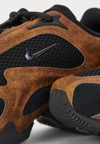 Les sneakers Nike présentent une combinaison de cuir texturé marron et de mesh noir, avec un accent de logo noir. La semelle est en caoutchouc noir avec un relief à motifs.