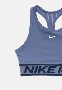 Nike sporta krūšturis gaiši zilā krāsā, ar platu pelēku joslu un tumši zilu tekstu. Tam ir sporta tipa aizmugure un gluda auduma tekstūra.