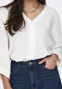 Biała koszula zapinana na guziki z teksturowanymi kropkami, z długimi rękawami i dekoltem w kształcie litery V. Połączona z ciemnoniebieskimi dżinsami. Widoczny prosty złoty naszyjnik.