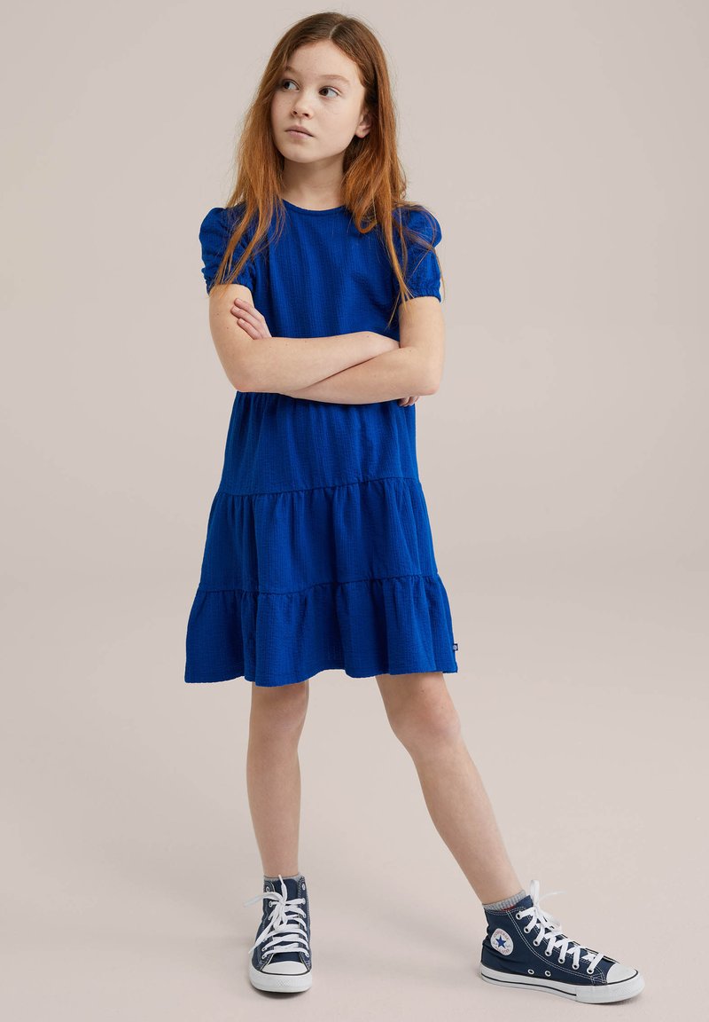 Blå bomulls klänning med flera lager och korta puffärmar, tillsammans med blå höga sneakers. Enkel design med texturerad tyg och rundad halsringning.