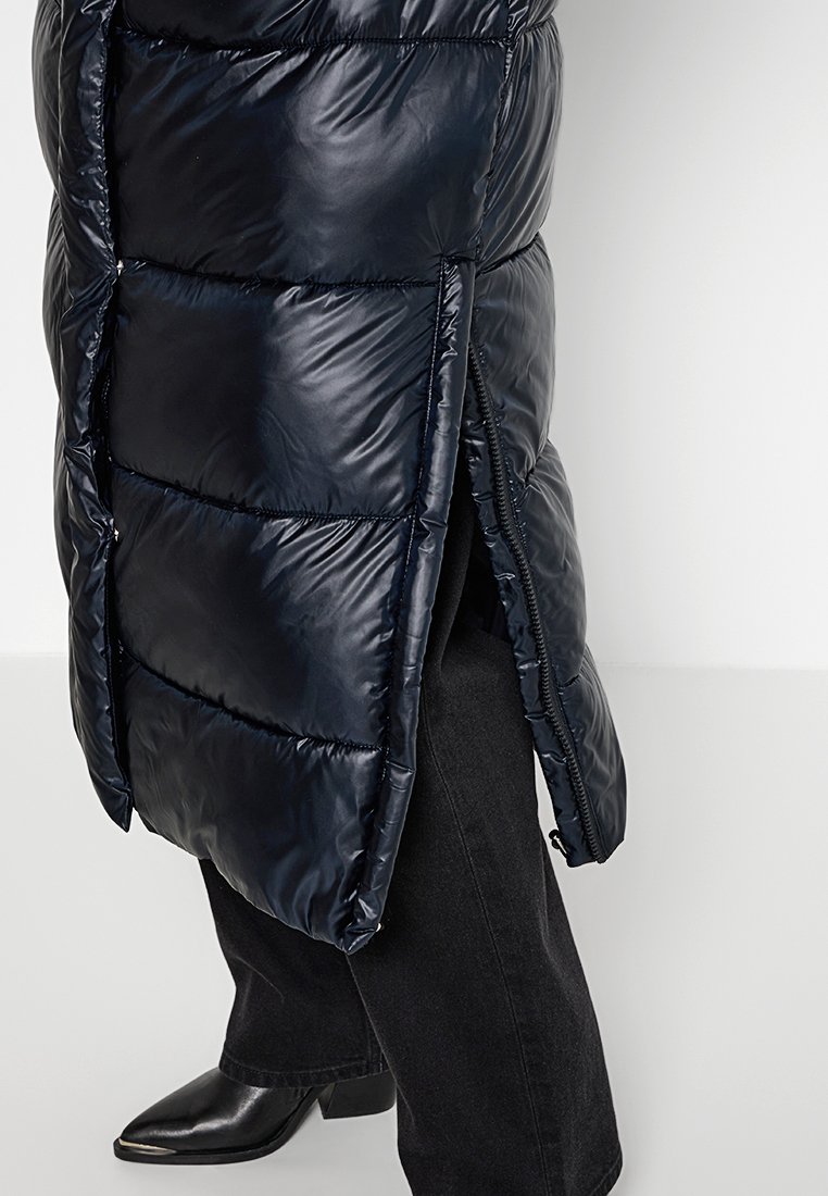 Doudoune marine à finition brillante, comportant des sections en quilting horizontal, des fentes latérales et une fermeture éclair sur toute la longueur.