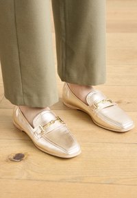 Mocassins en cuir doré avec un bout pointu et un accent décoratif en chaîne. Texture lisse, design bas, associées à un pantalon vert olive.