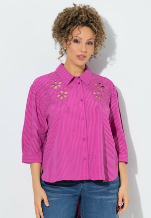 Mujer con cabello rizado lleva una blusa magenta abotonada con recortes florales y vaqueros azules, de pie frente a un fondo blanco.