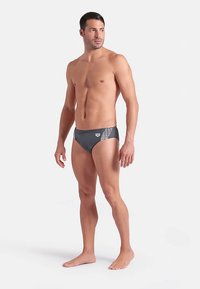 Bañador tipo slip para hombre en tela gris oscuro, con un diseño de malla en los costados, corte de tiro bajo y cintura elástica. Textura suave.