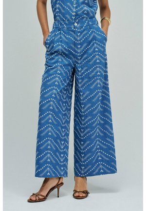 Pantalones anchos azules con patrones bordados en zigzag y florales en blanco, combinados con sandalias marrones de tacón alto y tiras, y una blusa a juego.