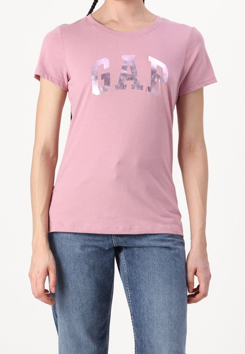 Femme portant un T-shirt rose clair à manches courtes avec des lettres argentées brillantes "GAP" et un jean bleu sur un fond blanc uni.
