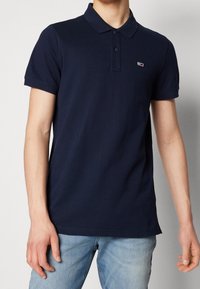 Man som bär en marinblå kortärmad polo med en liten röd och vit logotyp på bröstet samt ljusblå jeans.