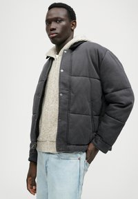 Geacă pufoasă gri cu un design matlasat, cu nasturi tip capsa și buzunare laterale, purtată peste un pulover tricotat deschis. Jeans de culoare bleu deschis, uzat.