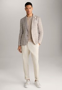 Blazer grigio chiaro con texture e due bottoni, abbinato a un maglione beige e pantaloni color crema, completato da sneakers bianche.