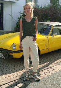 Jeune femme aux cheveux blonds portant un haut vert sans manches, un pantalon blanc et des baskets, se tient devant une voiture ancienne jaune.