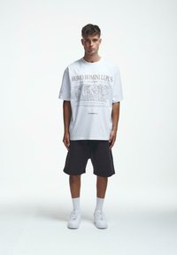 Balta per didelę t-shirt su grafiniu spaudiniu, kuriame yra tekstas "HOMO HOMINI LUPUS", derinama su tamsiomis šortais ir baltais sportbačiais. Lygi tekstūra.