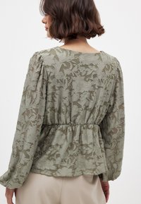 Ljus olivgrön blus med genomskinlig blommönster, rynkad midja, långa ärmar och vid halsringning. Mjuk, strukturerad tyg.